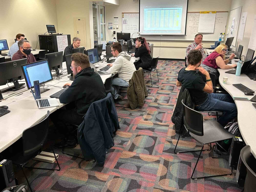 A classroom-style computer lab with nine adults seated at desks arranged along two curved rows, each working intently at desktop computers. A large projection screen at the front displays a spreadsheet, while the space is lit with overhead lights